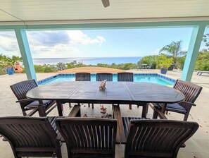 Un espace piscine extérieur avec une table et des chaises en bois, entouré d’une terrasse couverte offrant une vue sur l’océan.