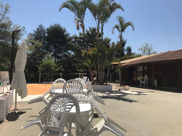 An outdoor seating area with white plastic chairs and tables, surrounded by palm trees and a building with a tiled roof.