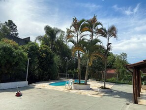 A swimming pool with a tiled deck, surrounded by trees and a building in the background. - Namaste house - Nature and good vibes: just 2km from the city center (Bragança Paulista)
