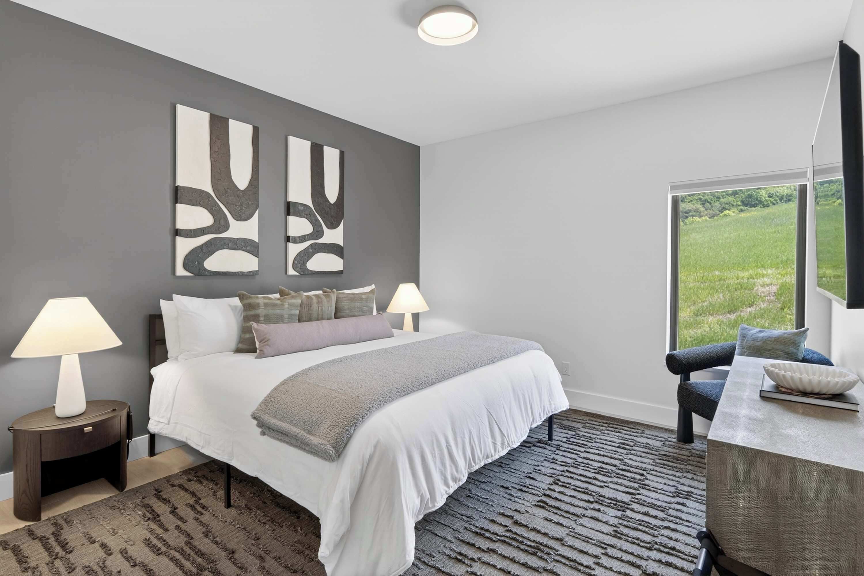 Une chambre moderne avec un grand lit, deux lampes de chevet, une vue sur un champ verdoyant par la fenêtre et des œuvres d’art abstraites accrochées au mur.
