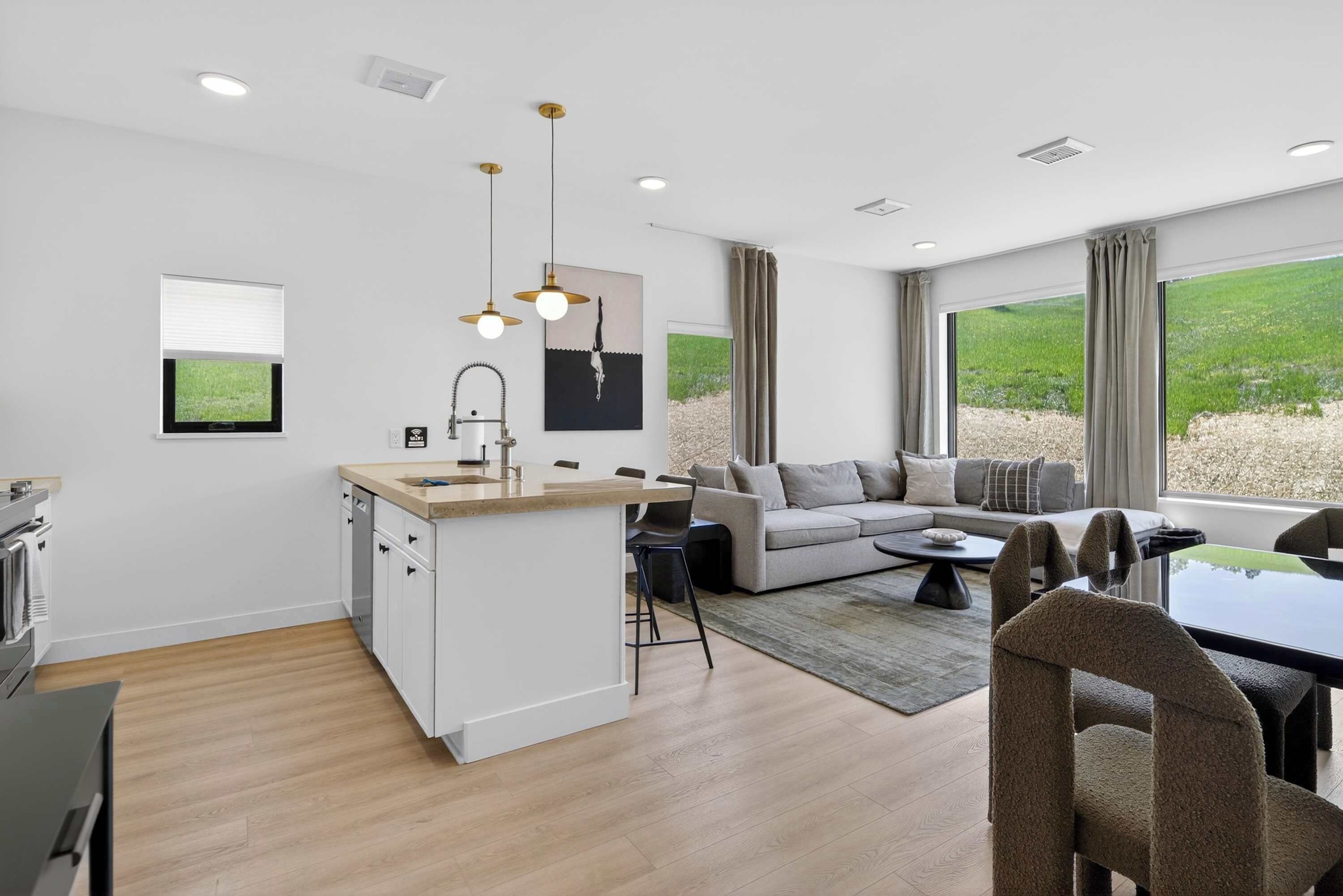 A modern kitchen with a central island, a dining area with a table and chairs, a sofa, and a large window with a view of greenery.