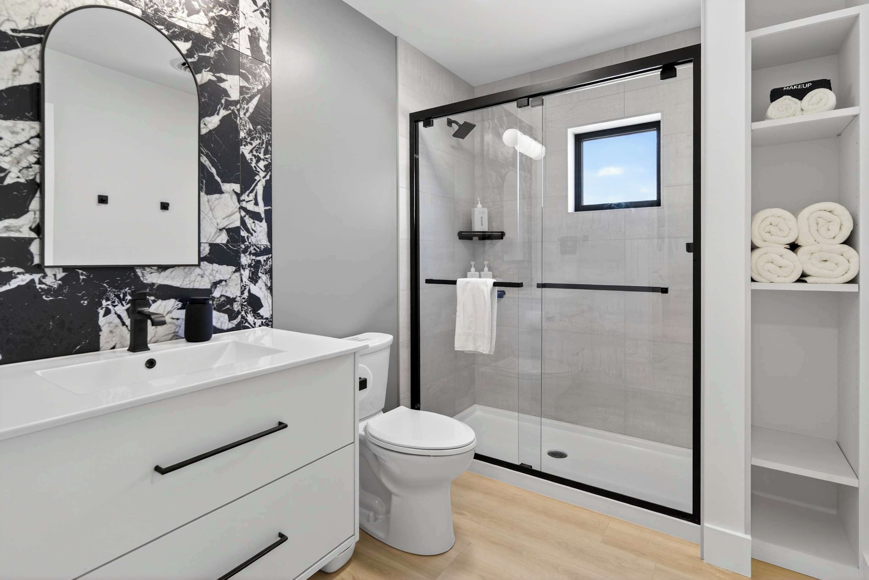 Ein modernes Badezimmer mit Marmorfliesen an der Wand, einem weißen Waschtisch mit schwarzem Wasserhahn, einer gläsernen Dusche und einem Regal mit aufgerollten Handtüchern.