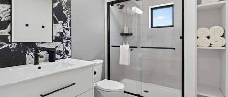 Ein modernes Badezimmer mit Marmorfliesen an der Wand, einem weißen Waschtisch mit schwarzem Wasserhahn, einer gläsernen Dusche und einem Regal mit aufgerollten Handtüchern.