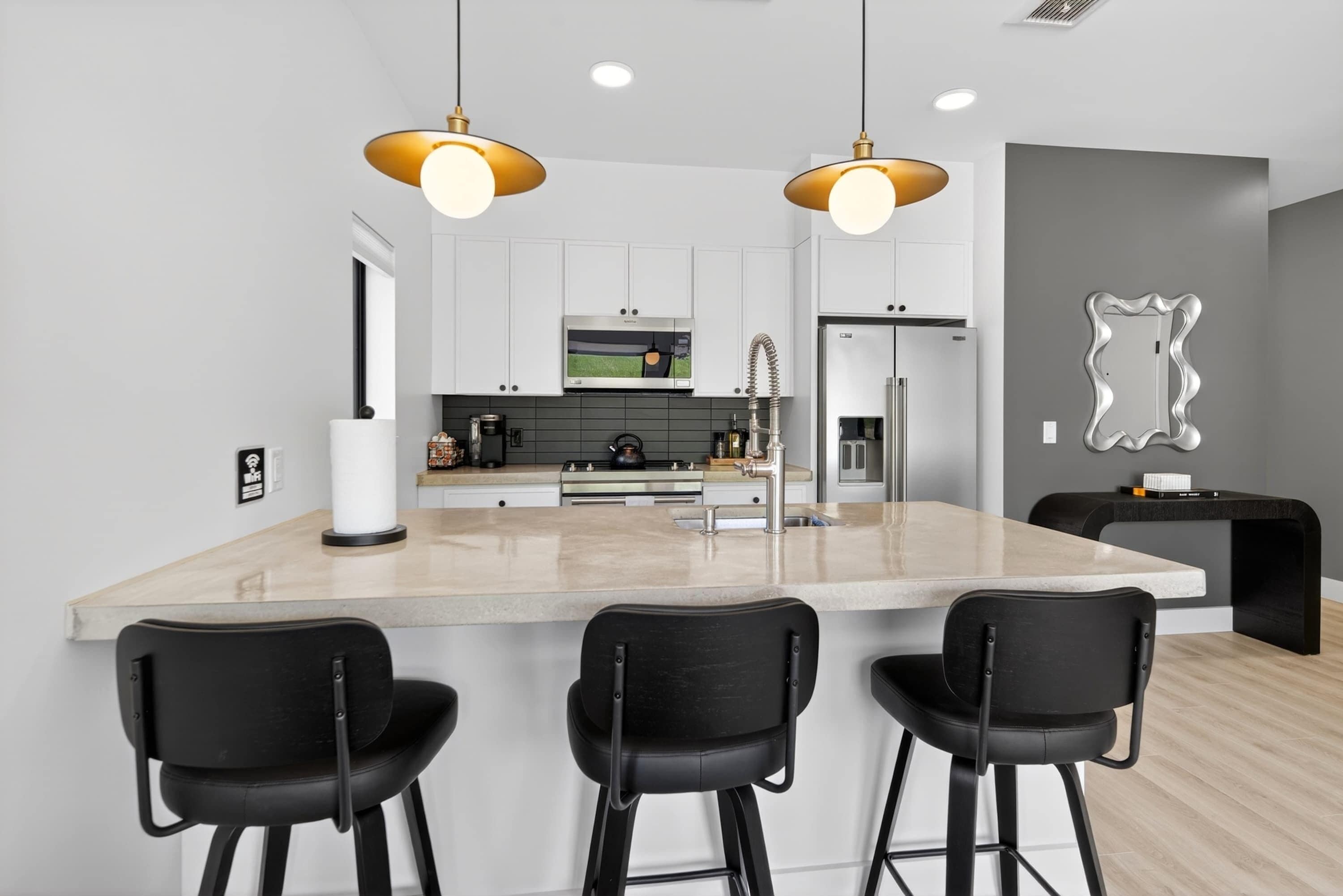 Une cuisine moderne dotée d’un îlot central, de tabourets de bar, d’appareils électroménagers en acier inoxydable et de luminaires suspendus.
