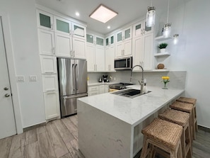 A modern kitchen with white cabinetry, stainless steel appliances, a marble island, and wicker stools. - Stunning Bay Views at Las Velas Village! (South Padre Island)