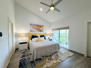 A bedroom with a bed, bedside tables, a ceiling fan, a painting, and a balcony with chairs. - Stunning Bay Views at Las Velas Village! (South Padre Island)