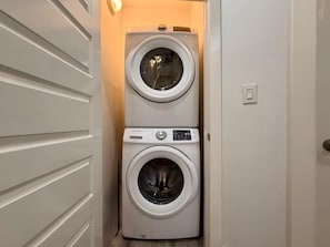 A laundry room with two stacked washing machines, a door with horizontal panels, and a light switch on the wall. - Stunning Bay Views at Las Velas Village! (South Padre Island)