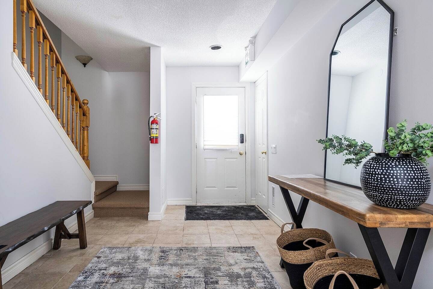 A hallway with a wooden staircase, a white door, a fire extinguisher, a large mirror, a wooden console table, and a rug.