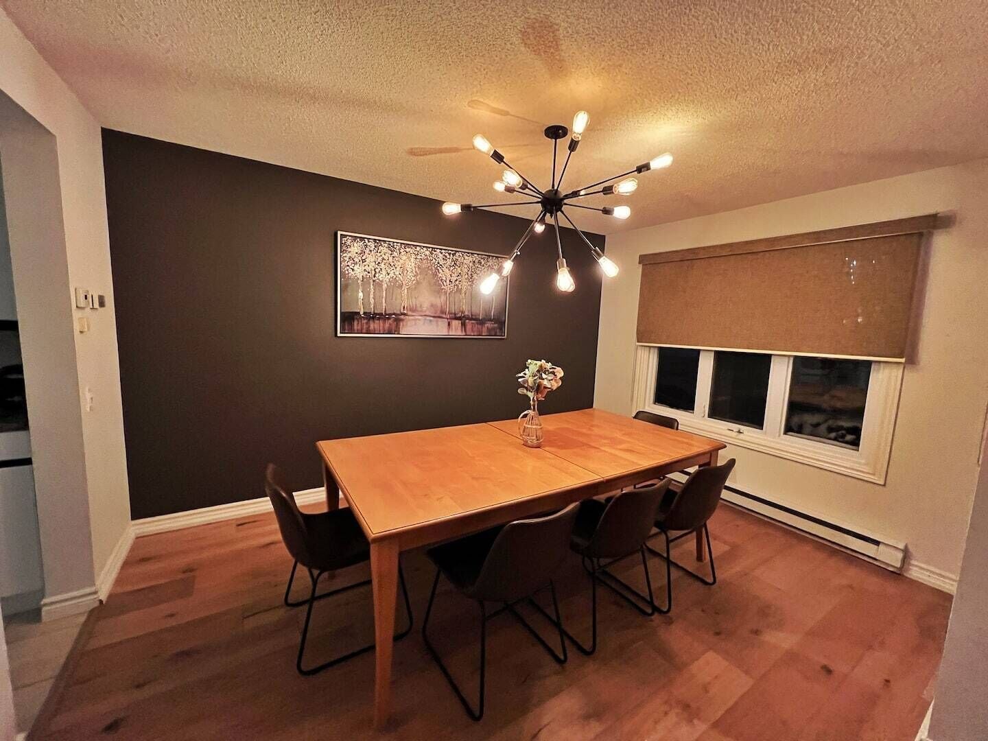 A dining area with a wooden table, black chairs, a modern ceiling light fixture, and a window with blinds.