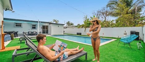 A poolside area with artificial grass, a lounge chair, and a person reading a magazine.