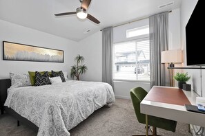 A hotel room with a bed, a desk, a chair, a ceiling fan, a window with curtains, a TV, and a plant. - Arcade • Covered Patio • Fire pit • Sleeps 10 (Nampa)
