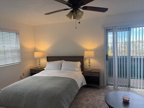 A hotel room with a bed, two bedside lamps, a ceiling fan, a window with blinds, and a view of the outdoors.