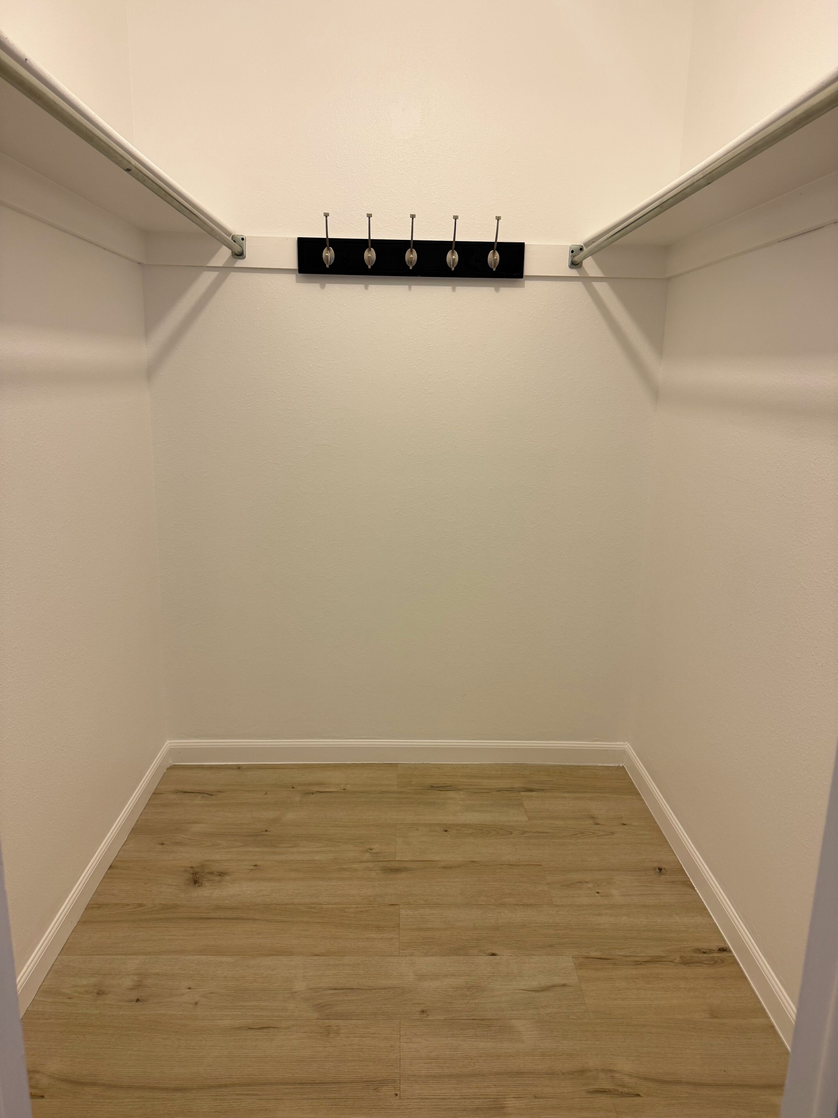 A corner closet with a wooden floor, white walls, and a black shelf with hooks.