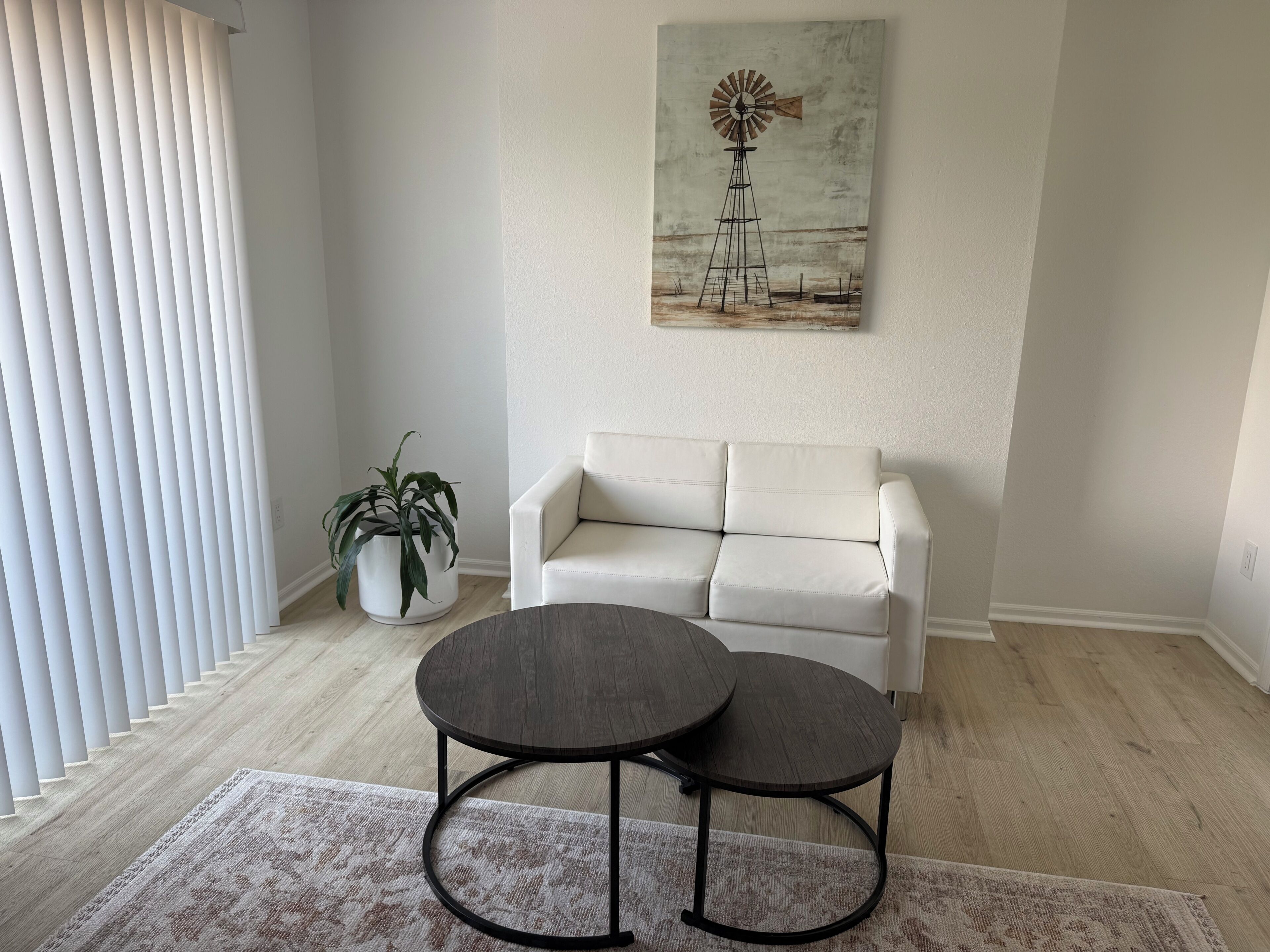 Un salon avec un canapé blanc, une table ronde accompagnée de deux chaises, une plante en pot et un tableau représentant un moulin à vent accroché au mur.