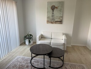A living room with a white sofa, a round table with two chairs, a potted plant, and a painting of a windmill on the wall.