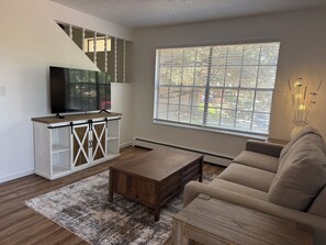 A living room with a TV, a sofa, a coffee table, and a large window with blinds.