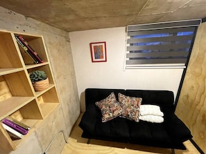 A small, cozy room with a black sofa, a wooden bookshelf, and a window with blinds.