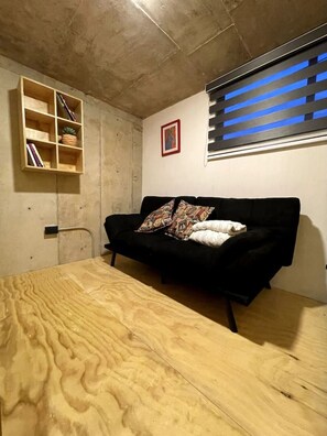 Une pièce avec un canapé noir, une table en bois, une bibliothèque et une fenêtre avec des stores.