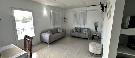 A living room with two sofas, a dining table with chairs, a small round ottoman, and a TV on a stand.