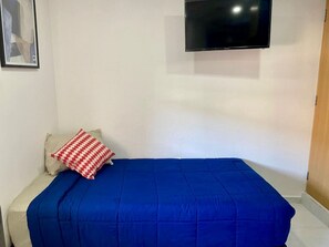 A single bed with a blue bedspread and a red and white patterned pillow, a flat-screen TV mounted on the wall, and a wooden door to the right. - Private space in Alameda W/ Kitchenette & bathroom (Santiago de Querétaro)