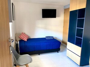 Ein kleiner, kompakter Raum mit Bett, Sessel, Wandfernseher und Kleiderschrank mit Ablagen.