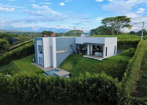Ein modernes, zweistöckiges Haus mit Swimmingpool, umgeben von üppiger Vegetation und Hügeln im Hintergrund.