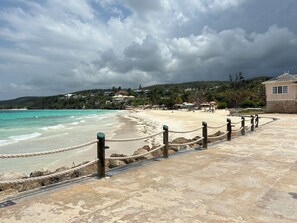 Ein Strand mit einer gepflasterten Promenade, einer Seilbarriere und einem Gebäude mit steinverkleideter Fassade.
