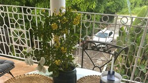 Una mesa redonda de cristal con individuales tejidos, una planta en maceta con flores amarillas y un vaso de acero inoxidable en un balcón con barandilla de metal.