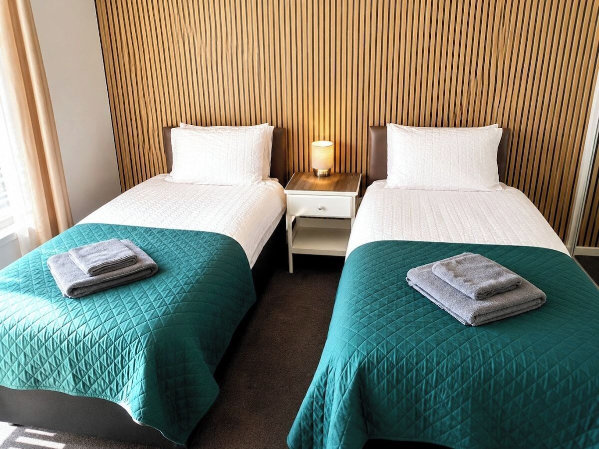 A hotel room with two beds, each with a teal quilted bedspread and a folded towel on top. There is a nightstand with a lamp between the beds.