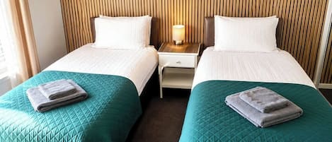 A hotel room with two beds, each with a teal quilted bedspread and a folded towel on top. There is a nightstand with a lamp between the beds.