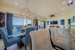 A modern living room with a glass dining table, blue chairs, a ceiling fan, and a view of the ocean. - 4BR Oceanfront Home | Incredible Views & Private Pools (Isla Mujeres)