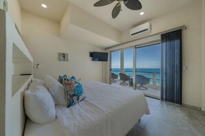 Ein Schlafzimmer mit einem Bett, einem Flachbildfernseher, einem Deckenventilator und Meerblick durch eine Glasschiebetür.