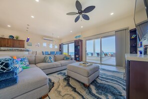 A spacious living area with a large sofa, an ottoman, and a dining table with chairs. There is a ceiling fan, a TV, and a view of the ocean through large sliding doors. - 4BR Oceanfront Home | Incredible Views & Private Pools (Isla Mujeres)