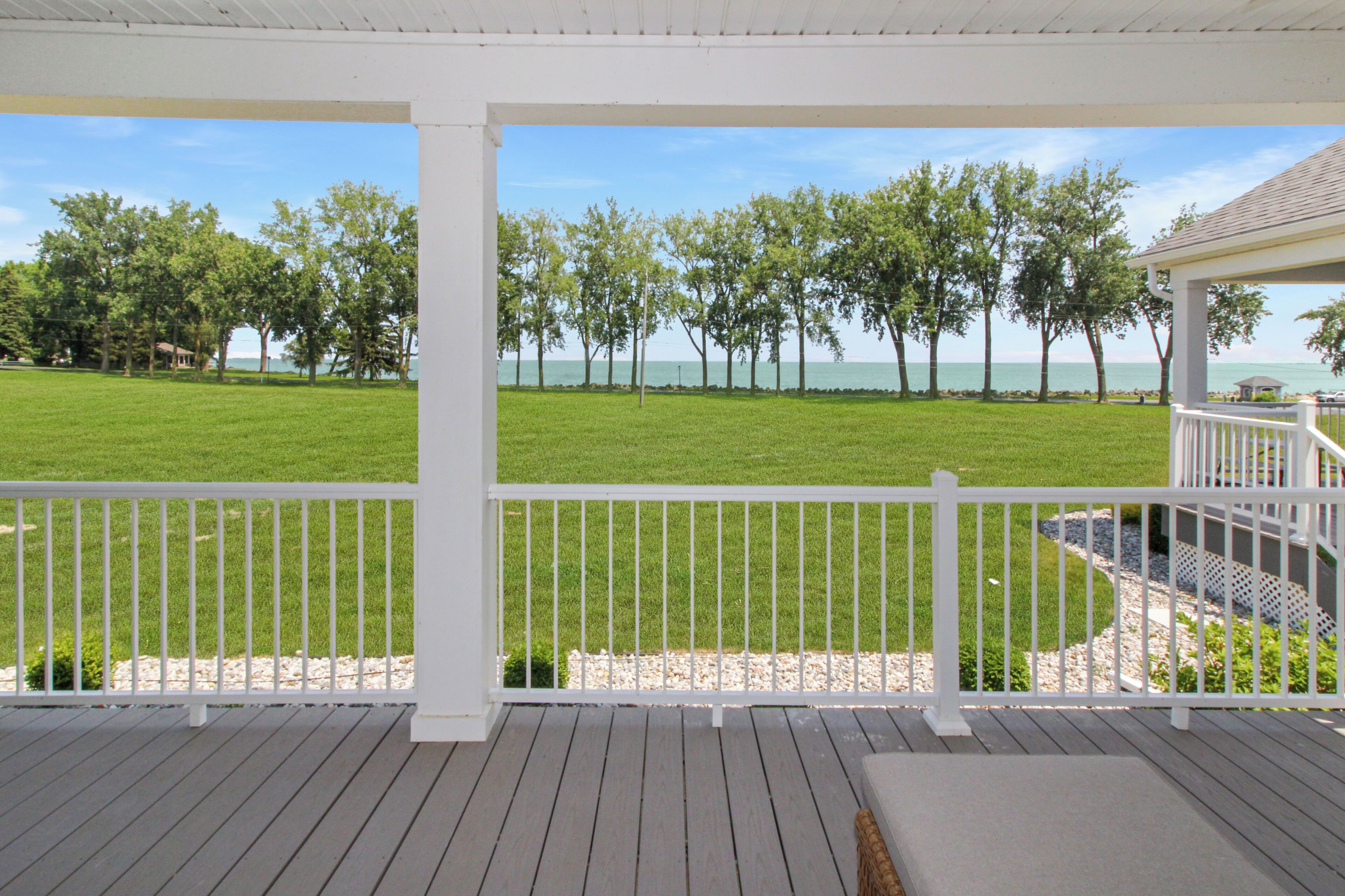 A covered outdoor area with a wooden deck, white railing, and a view of a grassy field and trees.