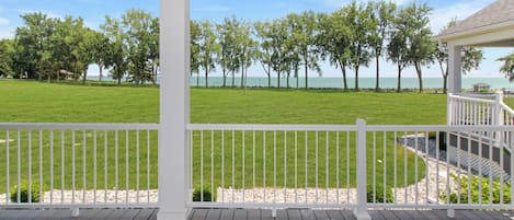 A covered outdoor area with a wooden deck, white railing, and a view of a grassy field and trees.
