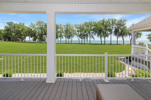 A covered outdoor area with a wooden deck, white railing, and a view of a grassy field and trees.
