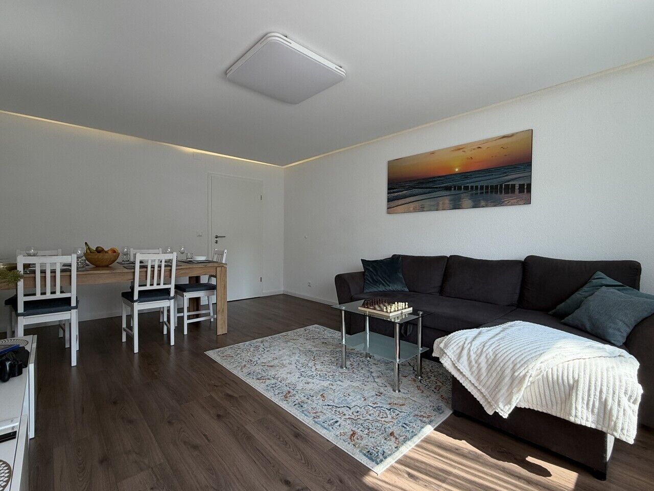 Ein modernes Wohnzimmer mit einem dunklen Sofa, einem Schachtisch und einem Gemälde, das einen Sonnenuntergang zeigt.