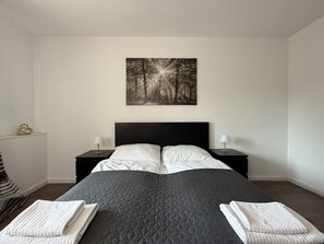 Een net opgemaakt bed met een gewatteerde dekbedovertrek, twee nachtkastjes met lampen en een aan de muur hangend beeld van een boslandschap.