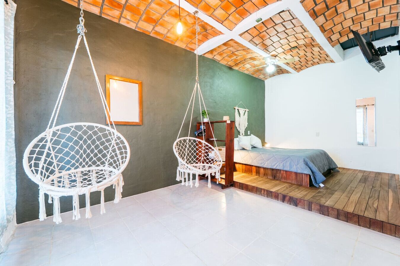 A bedroom with a bed, two hanging chairs, a wooden platform, and a brick ceiling.