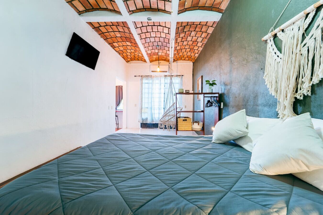 A bedroom with a bed, a macrame wall hanging, and a brick ceiling.