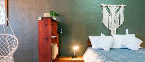 Een slaapkamer met een bed, een macramé wanddecoratie, een houten plankje met een plant, en een hangstoel.