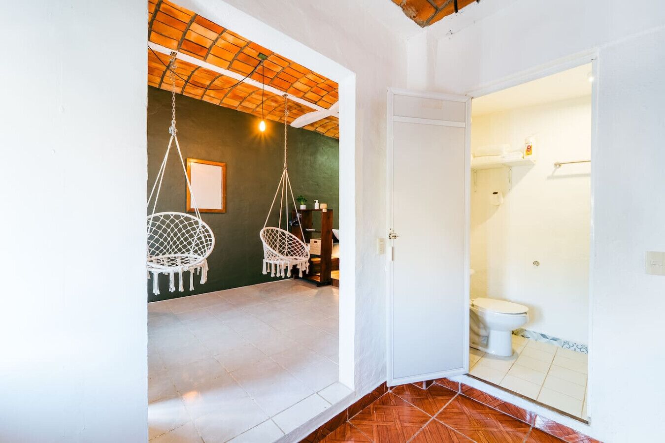 Un pasillo que conduce a un baño con inodoro y ducha. En la habitación contigua hay dos sillones colgantes blancos.