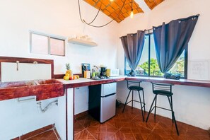 Une cuisine compacte équipée d’un évier, d’un four à micro-ondes et d’un petit réfrigérateur. On y trouve deux tabourets de bar et une fenêtre avec des rideaux.