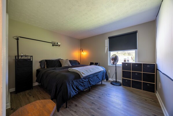 Un dormitorio con cama, escritorio, silla, cómoda, lámpara de pie y ventana con persianas.