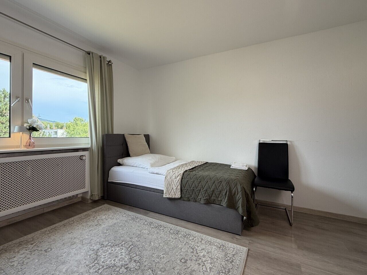 Ein Schlafzimmer mit Bett, Nachttisch, Stuhl, Heizkörper und einem fenster mit Vorhängen.