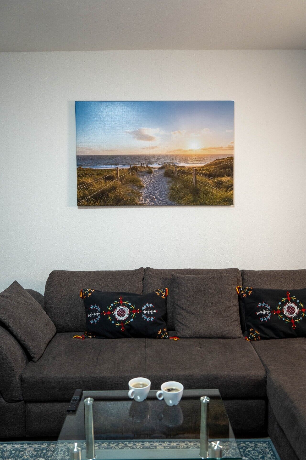 Ein Wohnzimmer mit einem großen Landschaftsbild, einer dunklen Couch mit aufgestickten Kissen und einem Glastisch mit zwei Tassen.