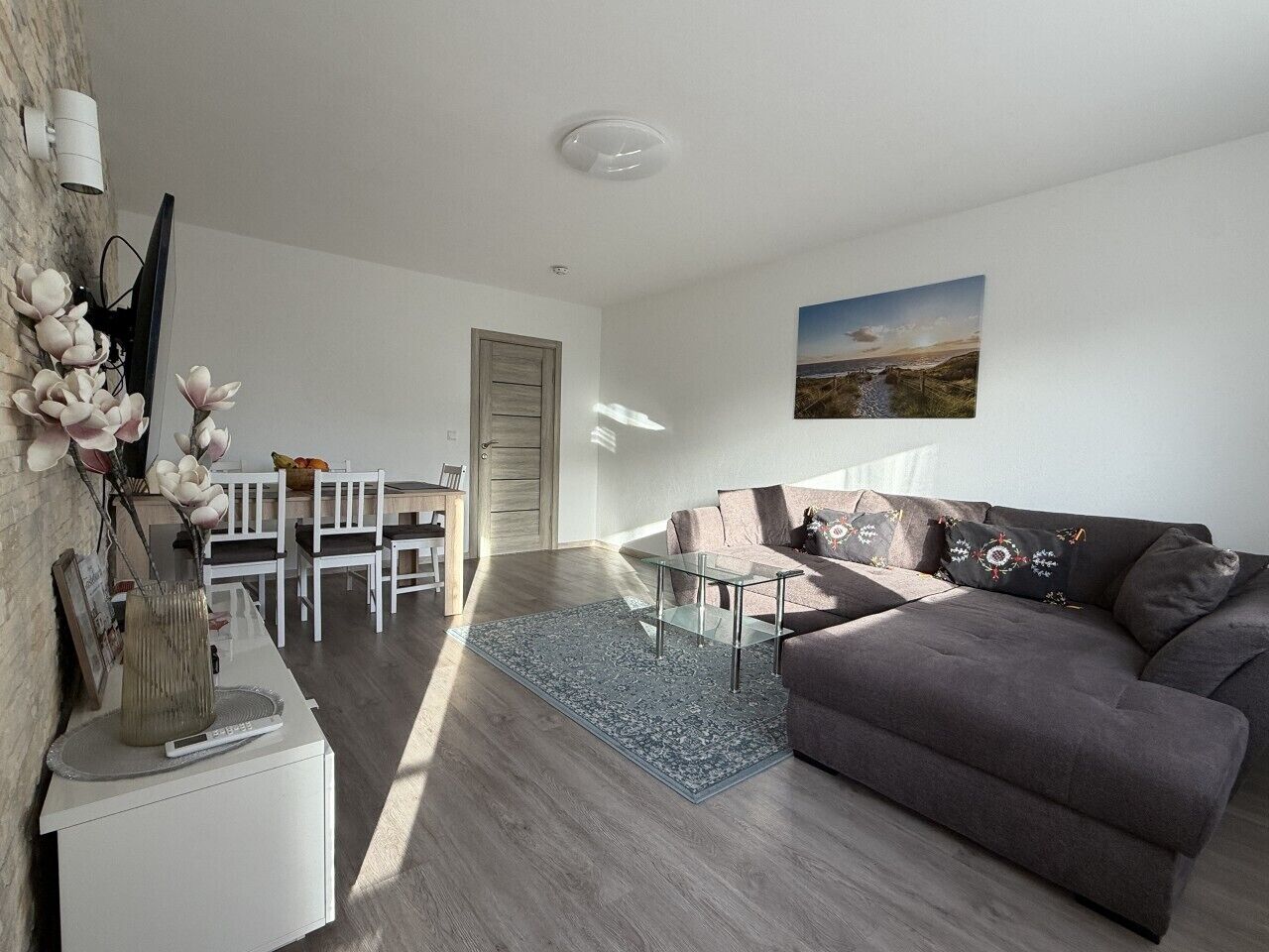 Ein modernes Wohnzimmer mit grauem Sofa, einem Glastisch und einem Essbereich mit weißen Stühlen.