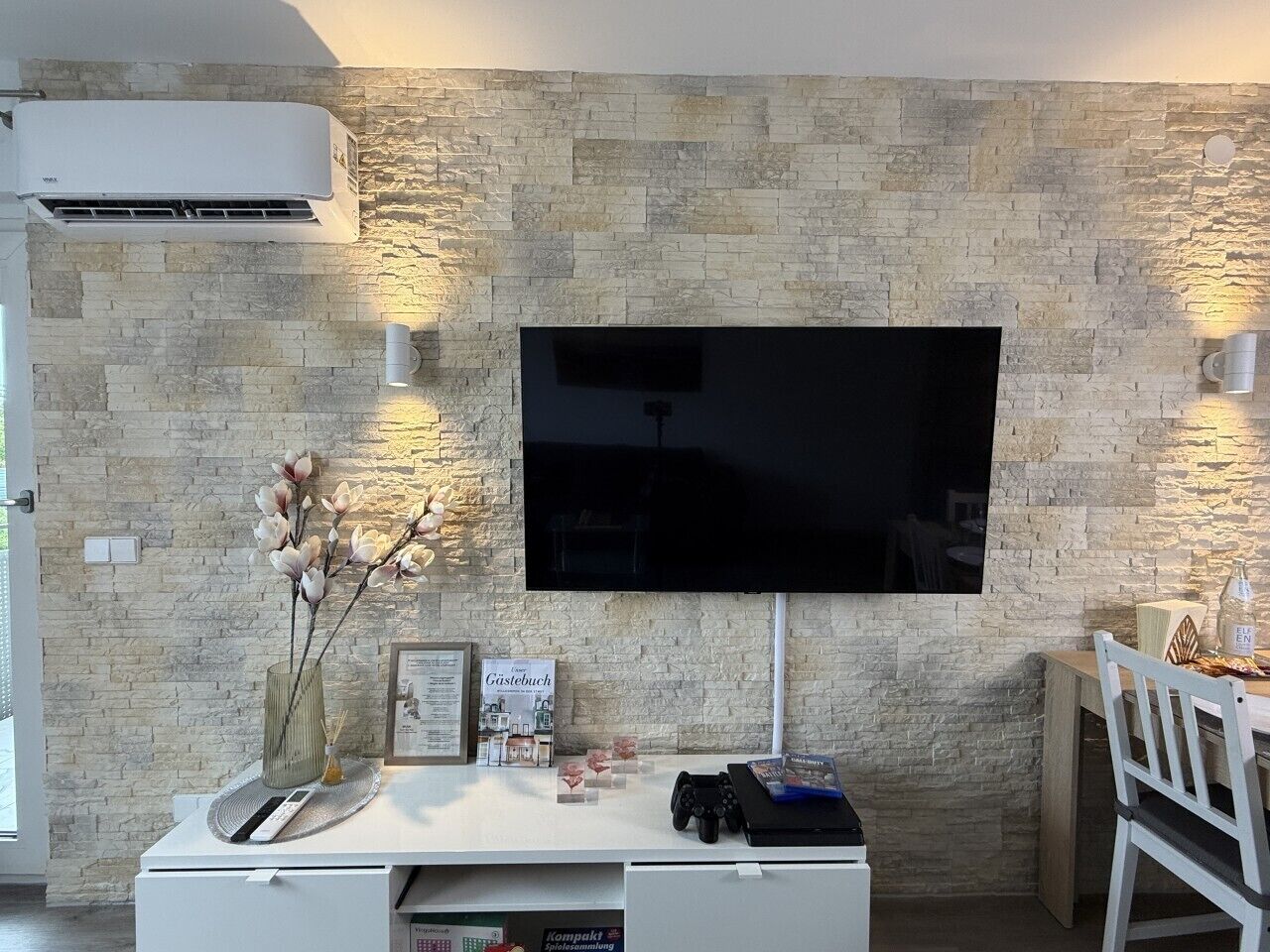 Ein modernes Wohnzimmer mit einem Flachbildfernseher an einer strukturierten Steinwand, einer weißen Unterhaltungsanlage inklusive Spielkonsole und einem kleinen Tisch mit einem Stuhl.