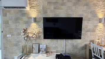 Ein modernes Wohnzimmer mit einem Flachbildfernseher an einer strukturierten Steinwand, einer weißen Unterhaltungsanlage inklusive Spielkonsole und einem kleinen Tisch mit einem Stuhl.