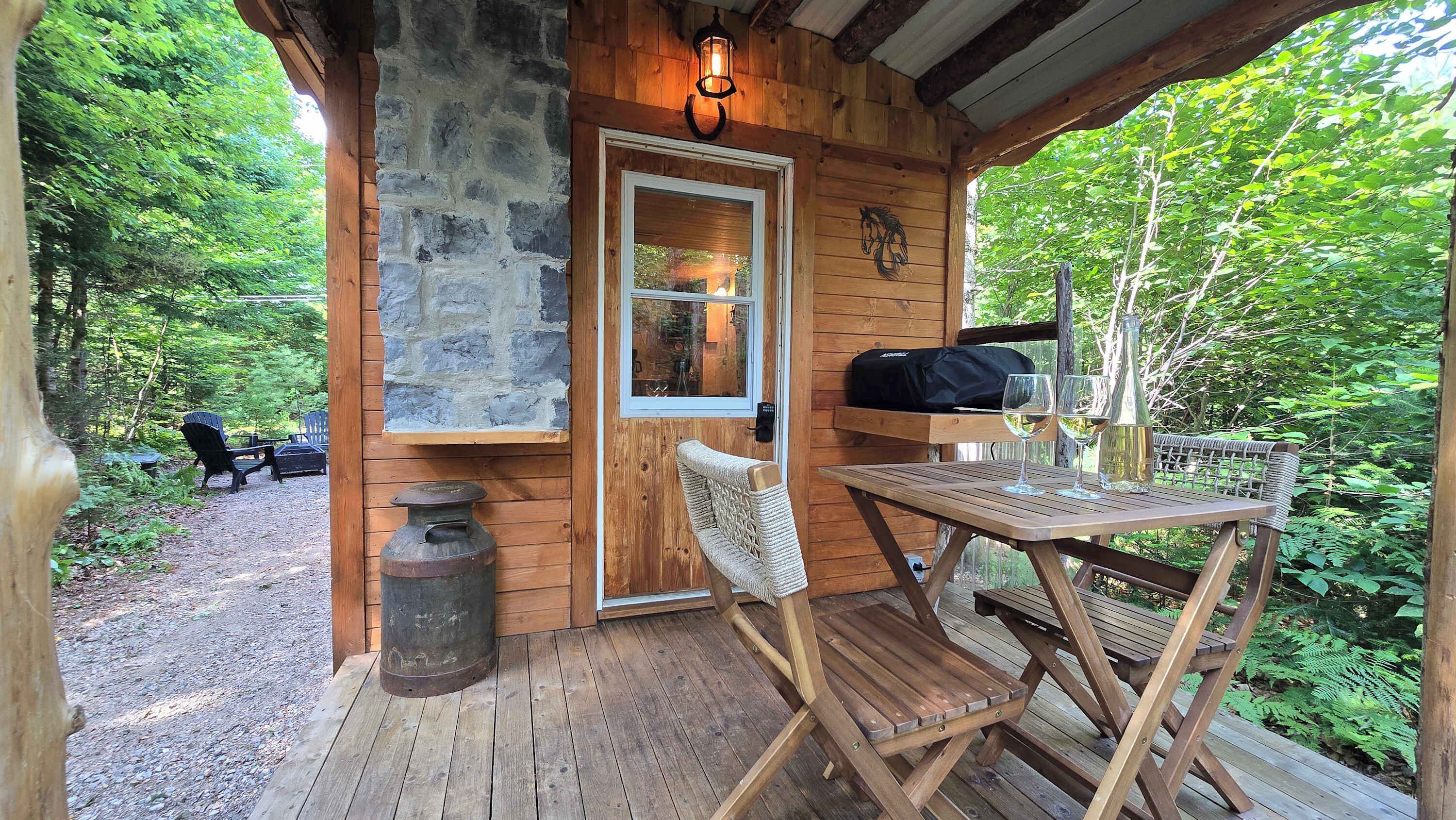 Une cabane en bois avec un mur en pierre, un banc, une table avec des verres et une bouteille, et une porte avec une fenêtre.